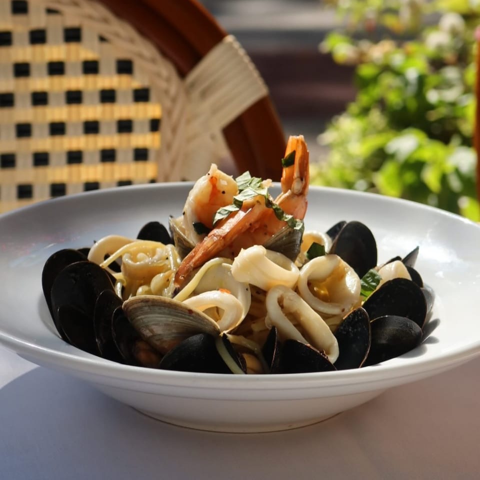 Bowl of seafood pasta topped with shrimp, mussels, clams, calamari, and herbs on an outdoor table.