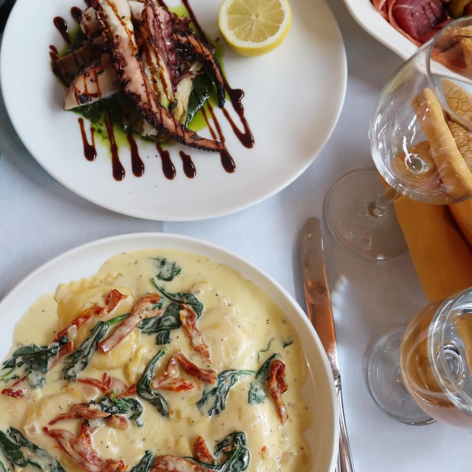Restaurant table featuring grilled octopus with lemon and balsamic drizzle, creamy pasta with spinach and tomatoes, wine, and appetizers.