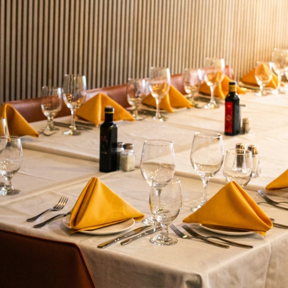 Elegant restaurant dining room with white tablecloths, golden folded napkins, wine glasses, cutlery, and bottles arranged for guests.
