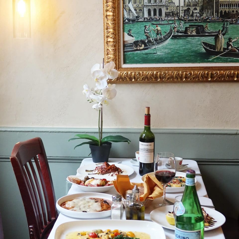 Italian restaurant table set with plated dishes, wine, water, white orchid, and a framed Venetian canal painting.