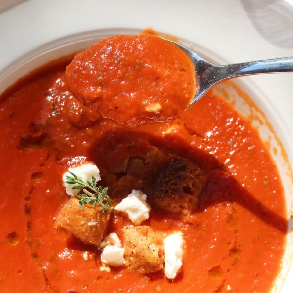 Spoonful of tomato sauce above a bowl of meatballs, croutons, feta, and fresh herbs.