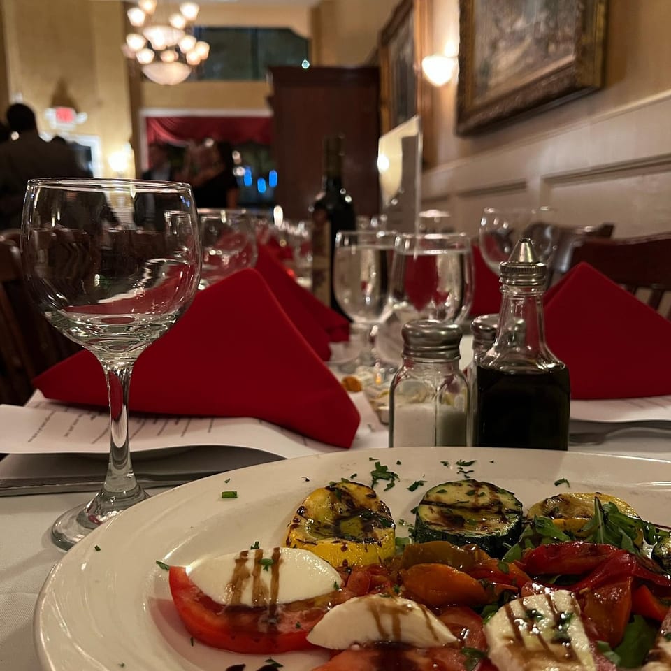 Elegant restaurant table with red napkins, wine glasses, condiments, and a plate of grilled vegetables, tomatoes, and fresh cheese.